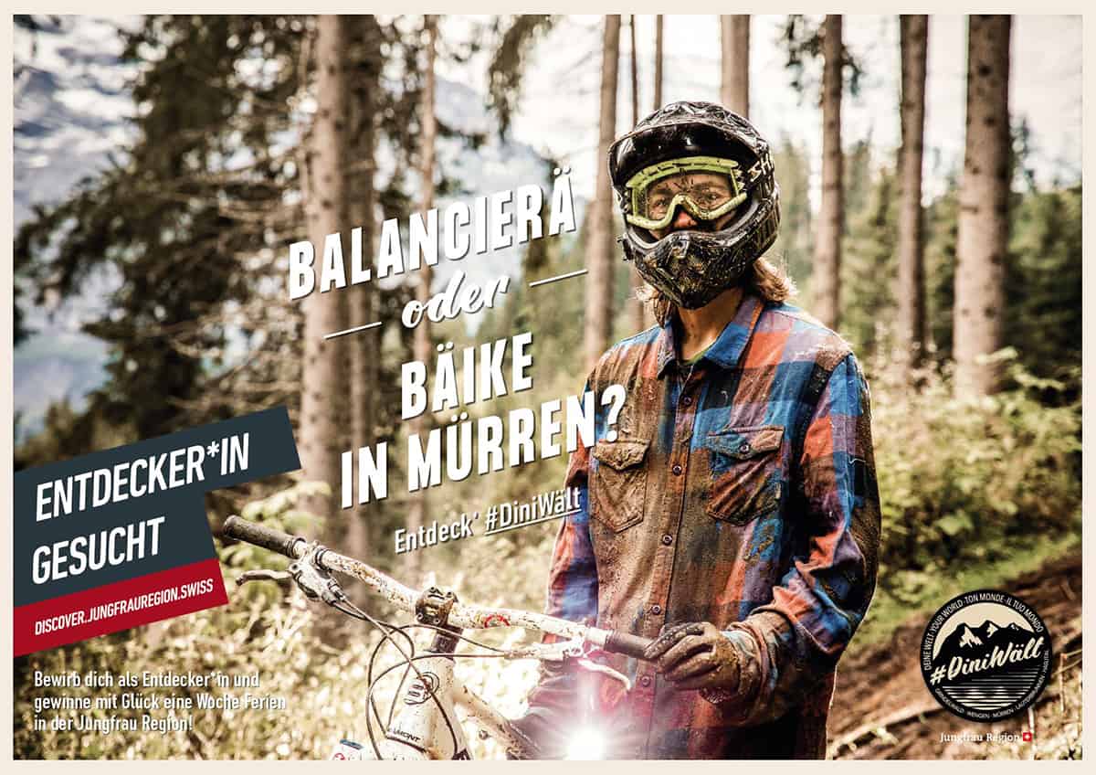 Auf einem Plakat der Kampagne #Dini Wält der Jungfrau-Region steht: «Balancierä oder bäike in Mürren? Entdecker*in gesucht.» Es zeigt einen dreckverschmierten Biker in Schutzausrüstung.