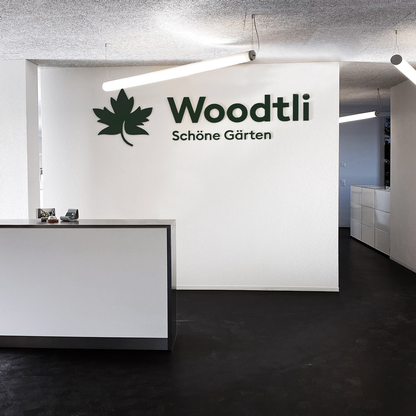 Am Empfang des Unternehmens sind an der weissen Wand im Hintergrund gross das Woodtli-Logo und der Slogan «Schöne Gärten» in Grün zu sehen.