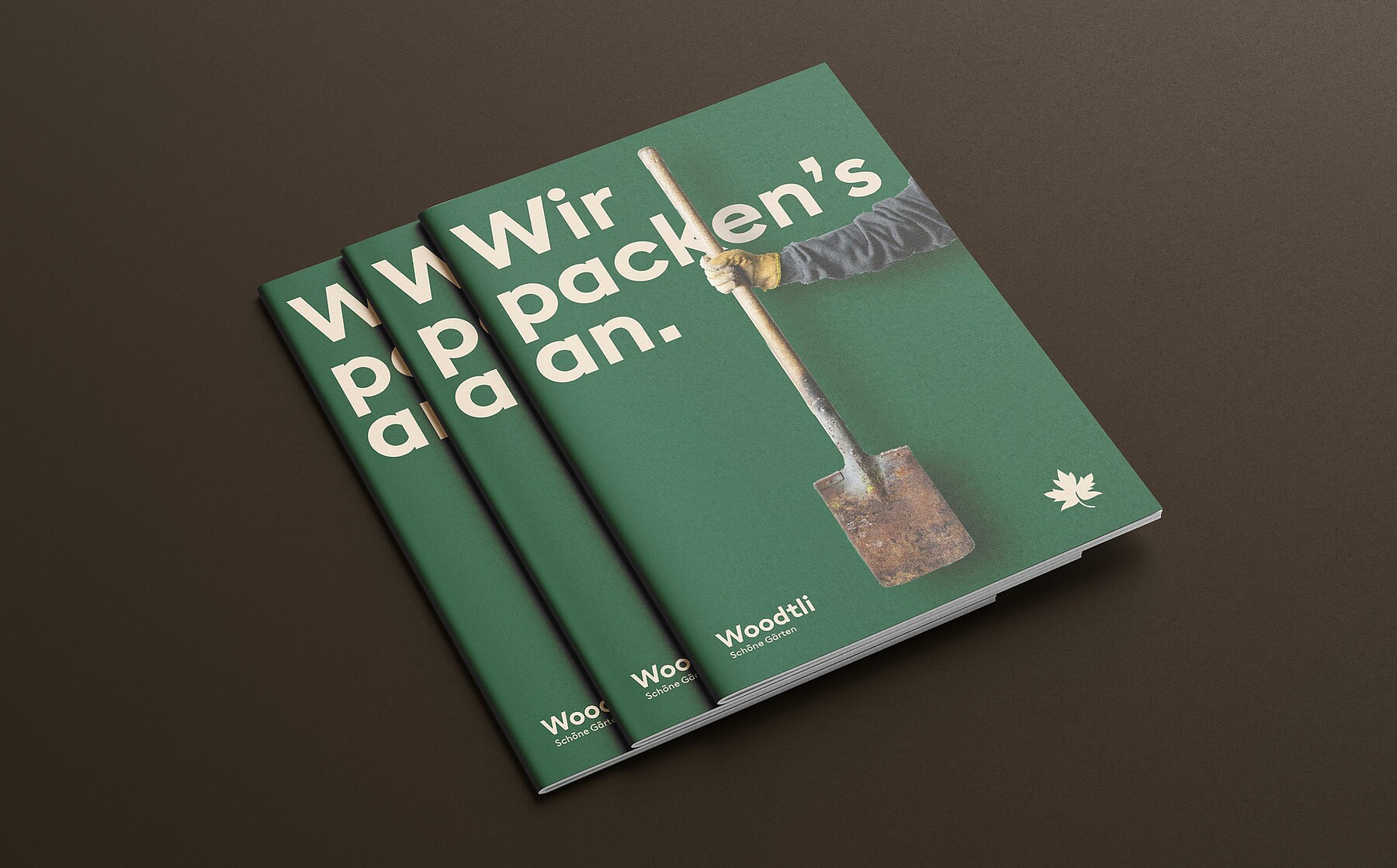 Mockups Design Auf dem grünen Cover einer Woodtli-Werbebroschüre steht: «Wir packen’s an.» Es zeigt eine Hand im Gartenhandschuh mit einer stark verwitterten Gartenschaufel.