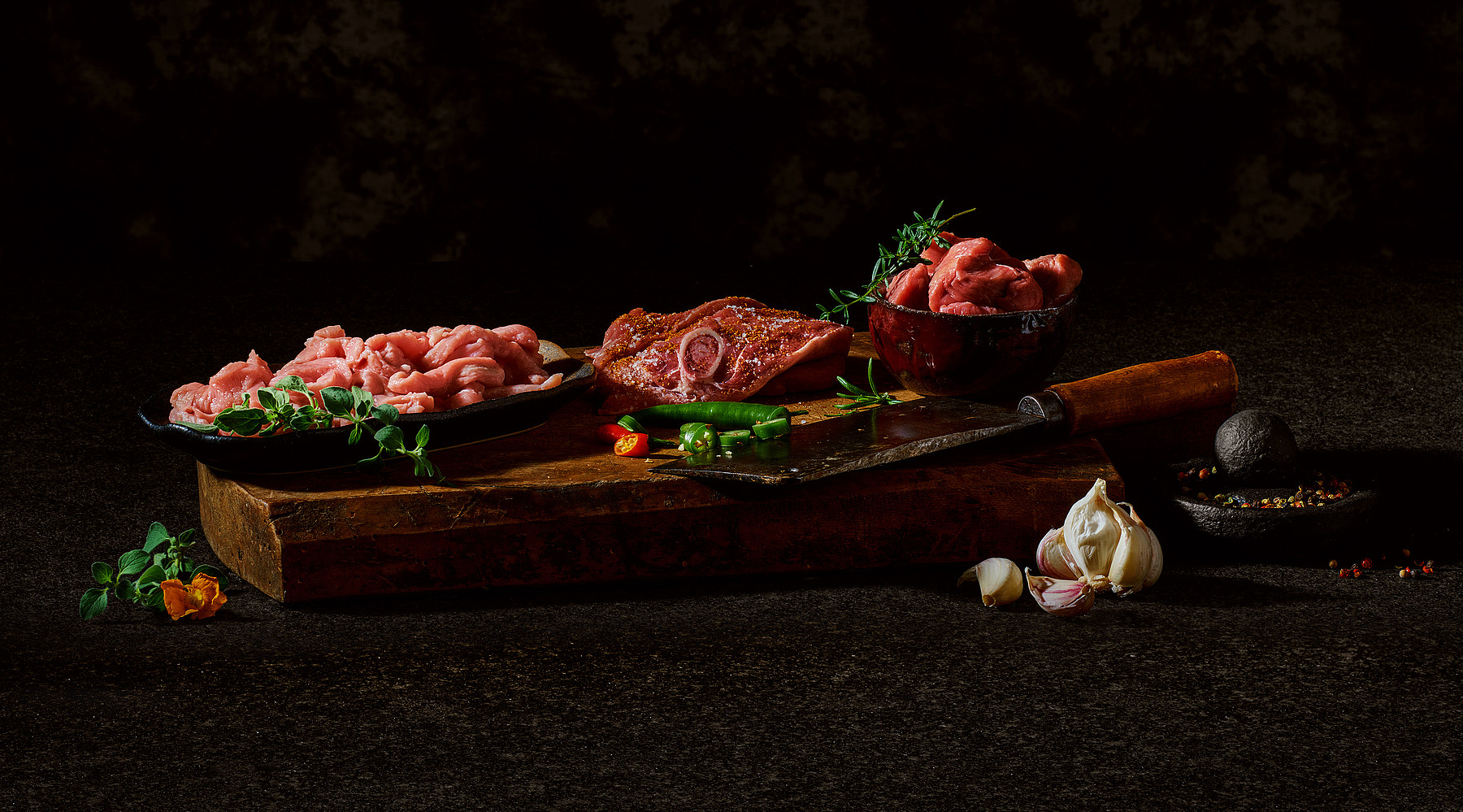 Rohe Fleischstücke präsentiert in Schälchen auf einem rustikalen Holzbrett, umgeben von Gewürzen
