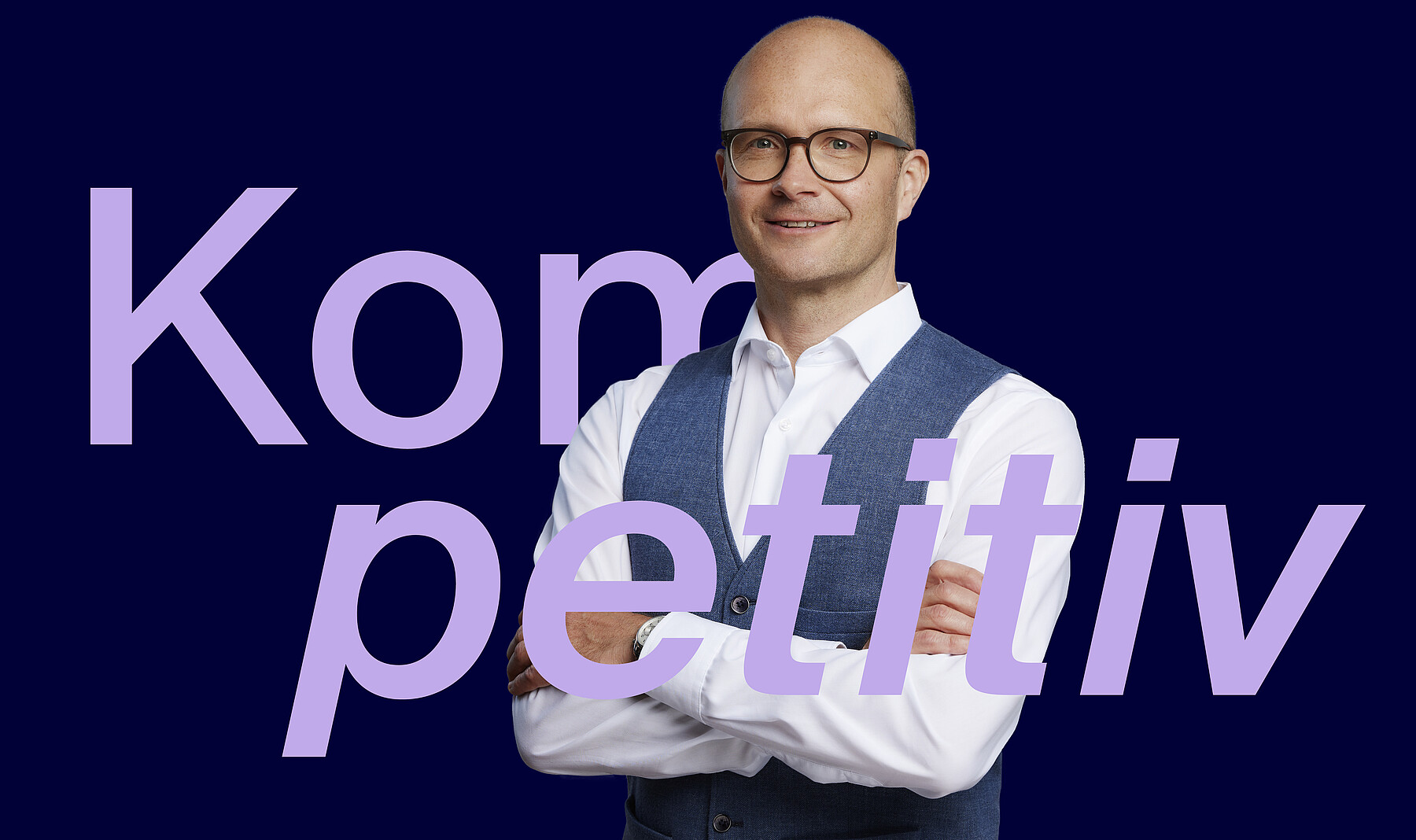 Mann mittleren Alters mit freundlichem, offenem Lächeln, kurzgeschorenen Haaren, Brille, weissem Hemd und jeansfarbenem Gilet, die Arme verschränkt. Bei ihm steht in grossen lilafarbenen Buchstaben das Wort Kompetitiv.