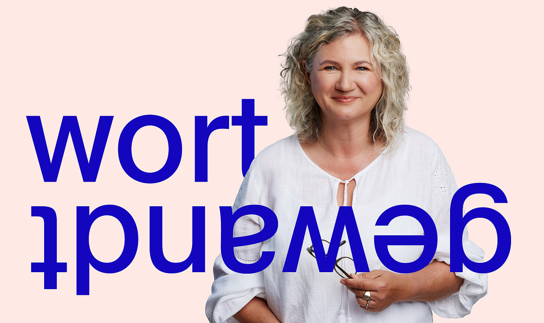 Frau mittleren Alters mit warmem Lächeln, blonden Locken, weisser Bluse und Lesebrille in der Hand. Bei ihr steht in grossen blauen Buchstaben das Wort Wortgewandt, die zweite Hälfte davon auf dem Kopf.