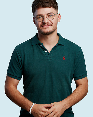 Junger Mann mit warmem Lächeln, dunklen Haaren, Brille und petrolfarbenem Poloshirt.