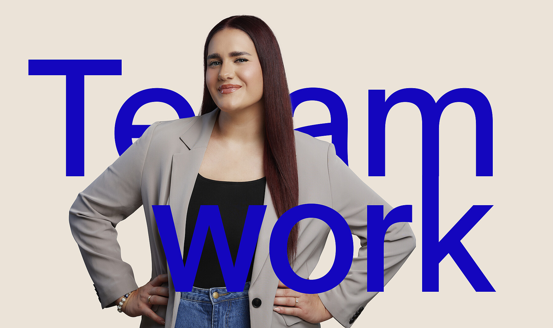 Junge Frau mit freundlichem, keckem Lächeln, langen dunklen Haaren und Blazer, die Arme auf die Hüfte gestützt. Bei ihr steht in grossen blauen Buchstaben das Wort Teamwork.